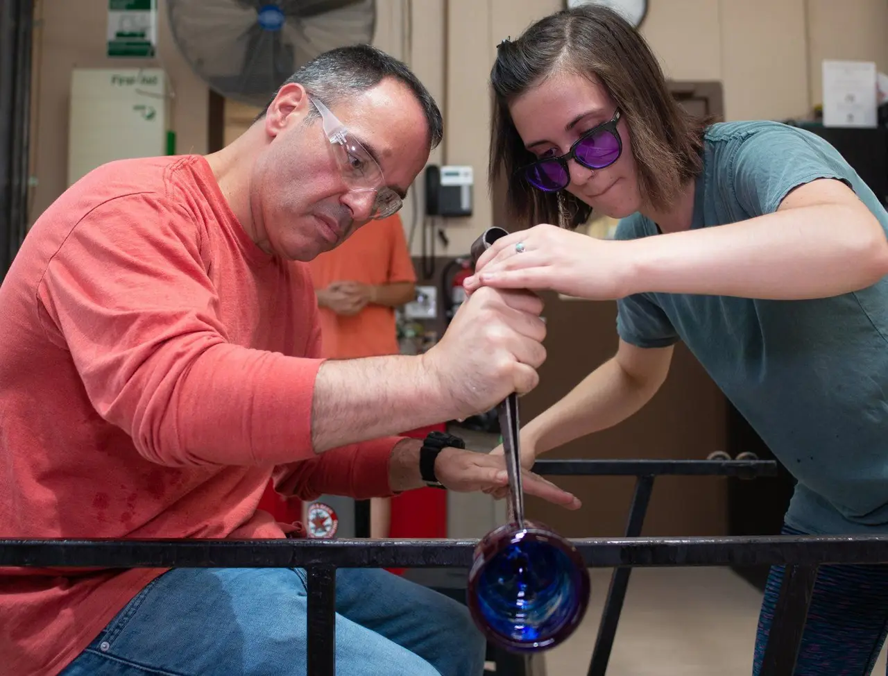 Instructor Hannah steadies the pipe as a student shapes a blue glass cup at the bench