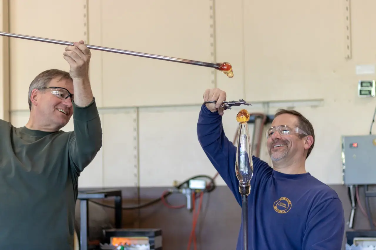 Two men laugh as they work a long pipe of glass together
