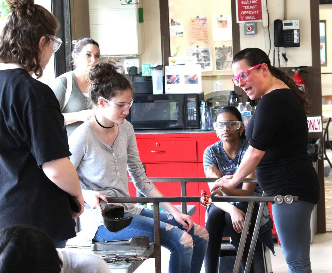Friends gather round the bench as an instructor helps one of them shape hot glass