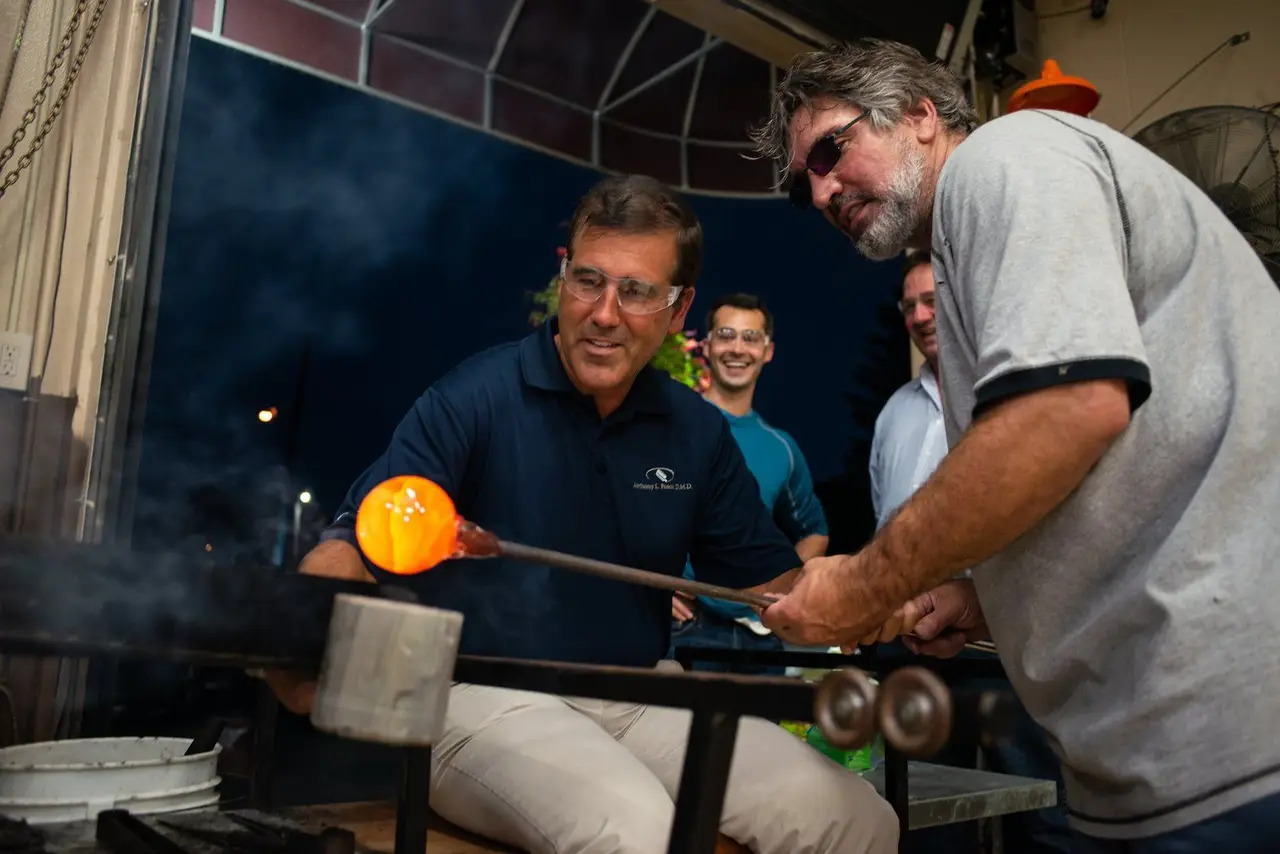 A visitor turns a glowing gather of molten glass at the glory hole while an instructor guides him
